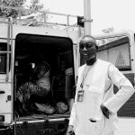 African journalist standing confidently beside a press van, captured in a street setting.
