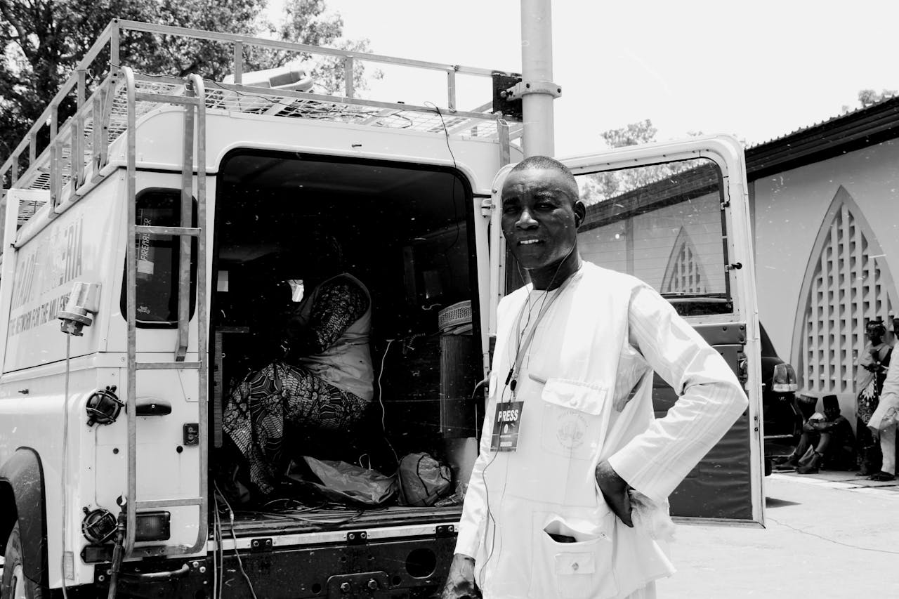 African journalist standing confidently beside a press van, captured in a street setting.