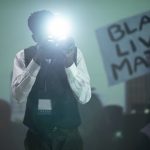 Photojournalist using camera flash at protest with Black Lives Matter sign.